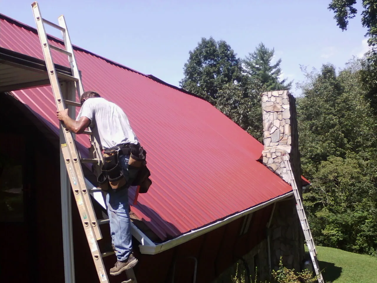 Roofer on a ladder beside a red metal roof and stone chimney — Soffit & Fascia Repair by Roofing Brothers in Kearny
